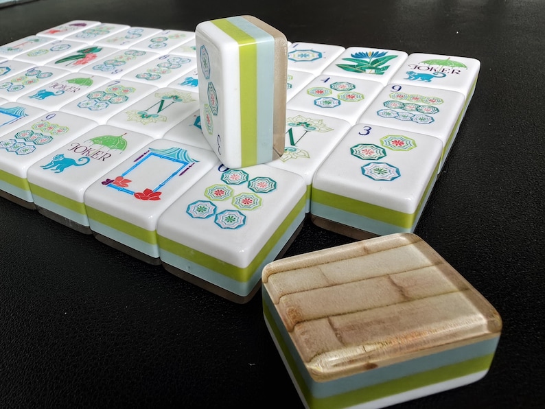 Puede incluir: Un juego de fichas de Mahjong rectangulares con caras blancas que presentan ilustraciones coloridas de flores, paraguas y la palabra JOKER. Las fichas tienen lados en capas de color verde, azul claro y madera, con una ficha vertical y otra plana.