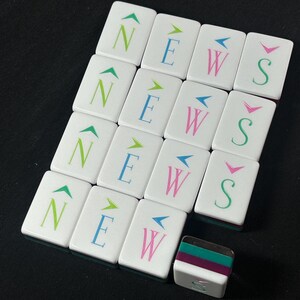 Op de afbeelding: Een set witte rechthoekige mahjongtegels gerangschikt in een raster, elk voorzien van een letter N, E, W of S in groen, blauw, roze of taling, bekroond met een richtingspijl. E&eacute;n tegel ligt horizontaal en toont een gelaagde groene en paarse zijkant.