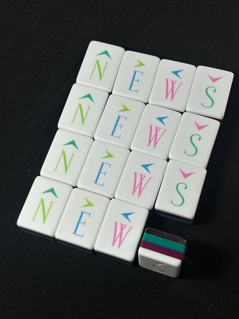 Op de afbeelding: Een set witte rechthoekige mahjongtegels gerangschikt in een raster, elk voorzien van een letter N, E, W of S in groen, blauw, roze of taling, bekroond met een richtingspijl. E&eacute;n tegel ligt horizontaal en toont een gelaagde groene en paarse zijkant.