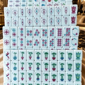 May include: A set of white Mahjong tiles arranged in rows and columns, wrapped in clear plastic. The tiles feature colorful engraved designs including Chinese characters, numbers, floral patterns, bamboo stalks, joker labels, and stylized dragon faces.