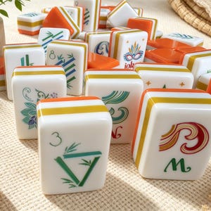K&ouml;nnte beinhalten: Eine Sammlung wei&szlig;er Mahjong-Spielsteine mit bunten grafischen Motiven, darunter Bambusstangen, Zahlen und eine Jokermaske. Die Spielsteine haben orangefarbene und gelbe horizontale Streifen an den Seiten und sind verstreut angeordnet.