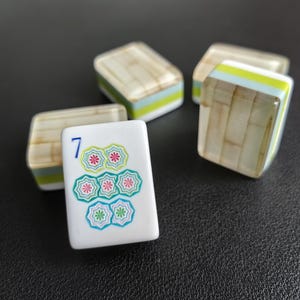 Puede incluir: Cuatro fichas de mahjong rectangulares con cara blanca y reverso con patr&oacute;n de bamb&uacute; marr&oacute;n claro. La ficha frontal muestra un n&uacute;mero 7 azul sobre un grupo de siete iconos con forma de flor en tonos verdes y rosas con centros blancos.