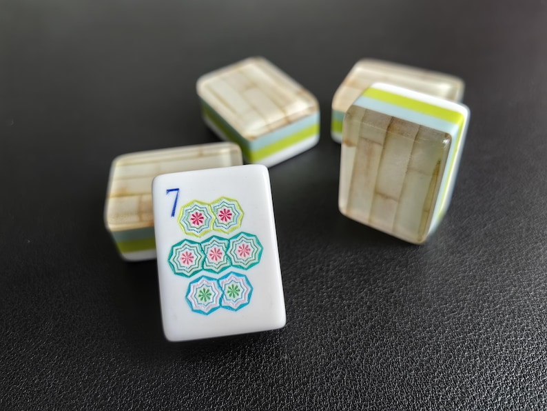 Puede incluir: Cuatro fichas de mahjong rectangulares con cara blanca y reverso con patr&oacute;n de bamb&uacute; marr&oacute;n claro. La ficha frontal muestra un n&uacute;mero 7 azul sobre un grupo de siete iconos con forma de flor en tonos verdes y rosas con centros blancos.