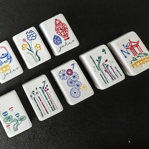 May include: Eight white rectangular Mahjong tiles featuring colorful illustrations including a teapot, flowers, a vase with the text jaker, a number 7, bamboo stalks, and a pagoda. Each tile displays detailed line art in blue, red, green, and yellow.
