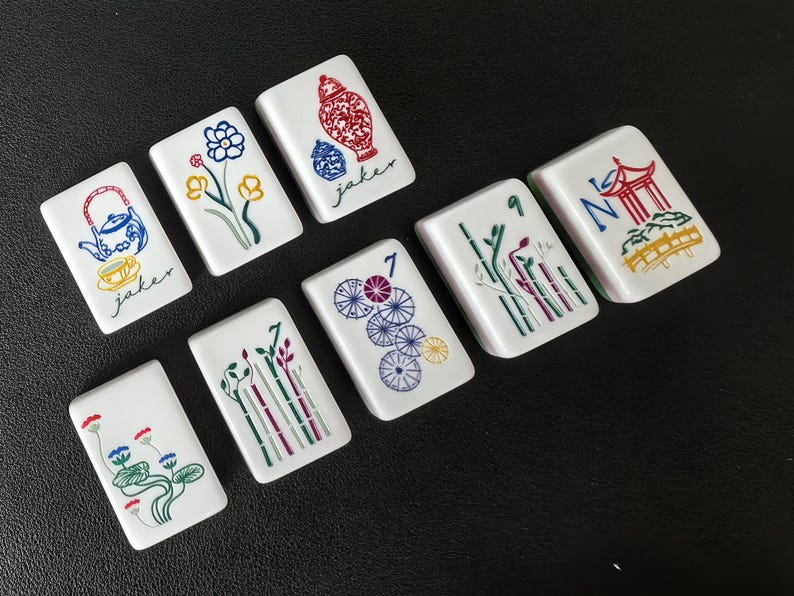 May include: Eight white rectangular Mahjong tiles featuring colorful illustrations including a teapot, flowers, a vase with the text jaker, a number 7, bamboo stalks, and a pagoda. Each tile displays detailed line art in blue, red, green, and yellow.
