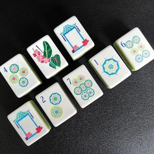 Puede incluir: Un conjunto de ocho fichas de mahjong rectangulares de color blanco con dise&ntilde;os impresos en color. Las fichas muestran varios patrones, incluyendo motivos florales azules y verdes, estructuras de pagodas estilizadas y n&uacute;meros en tinta azul.