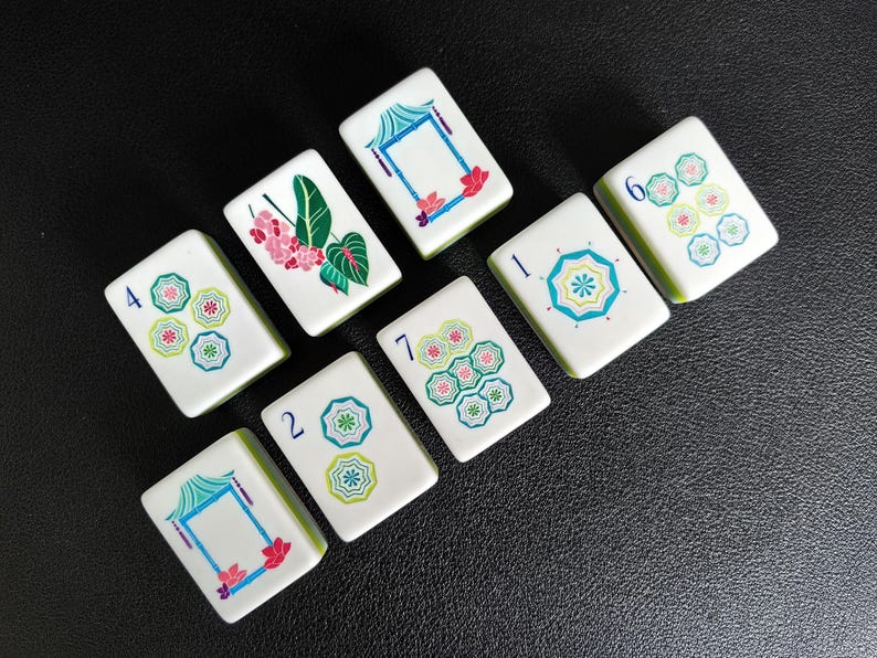Puede incluir: Un conjunto de ocho fichas de mahjong rectangulares de color blanco con dise&ntilde;os impresos en color. Las fichas muestran varios patrones, incluyendo motivos florales azules y verdes, estructuras de pagodas estilizadas y n&uacute;meros en tinta azul.