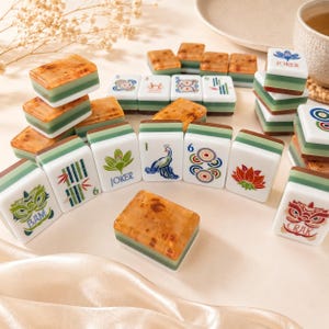 May include: A collection of Mahjong tiles featuring a mix of white faces with colorful illustrations of flowers, birds, and symbols, alongside brown marbled backs. Some tiles display text including JOKER, BAM, and CRAK, arranged in a scattered display.
