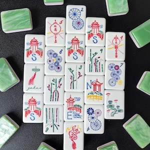 May include: A collection of rectangular mahjong tiles with white faces featuring colorful engraved illustrations of pagodas, bamboo, dragons, and floral patterns, alongside solid green marbled backs arranged in a scattered pattern.