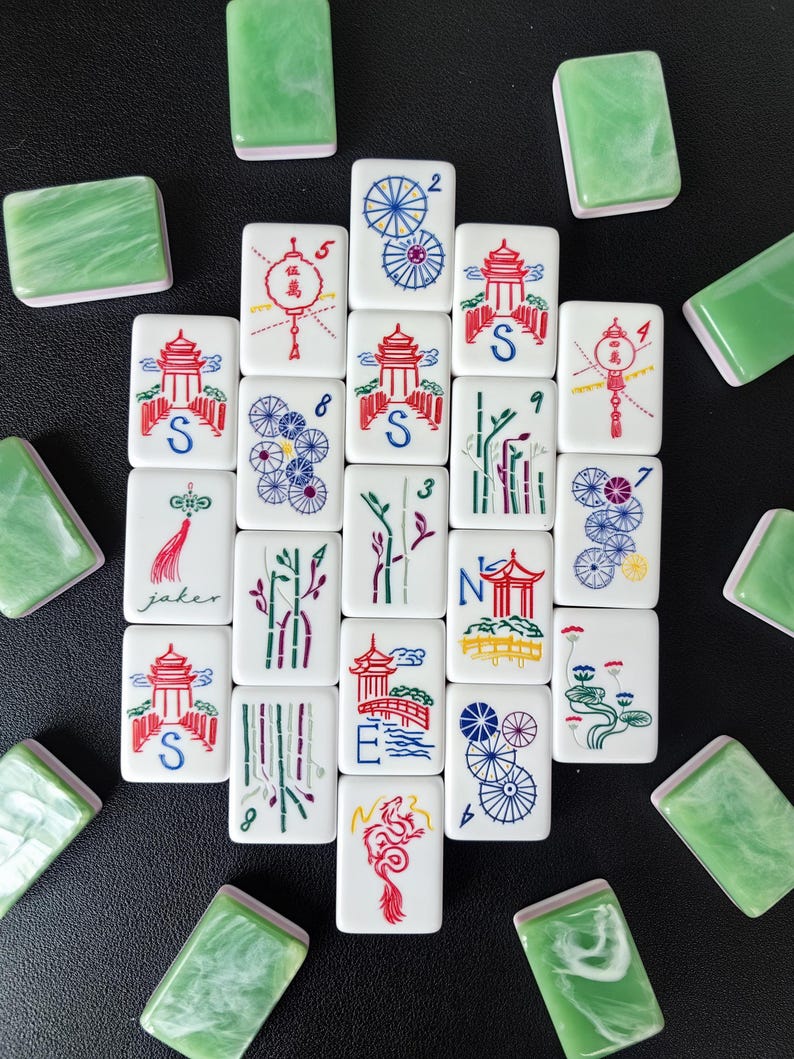 May include: A collection of rectangular mahjong tiles with white faces featuring colorful engraved illustrations of pagodas, bamboo, dragons, and floral patterns, alongside solid green marbled backs arranged in a scattered pattern.