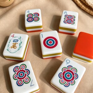 K&ouml;nnte beinhalten: Ein Set aus sieben rechteckigen Mahjong-Spielsteinen mit wei&szlig;en Oberfl&auml;chen und mehrfarbigen Seiten. Die Steine zeigen verschiedene Motive, darunter konzentrische Kreise, florale Muster, die Zahlen 4, 8, 1, 9 und das chinesische Schriftzeichen f&uuml;r zehntausend.