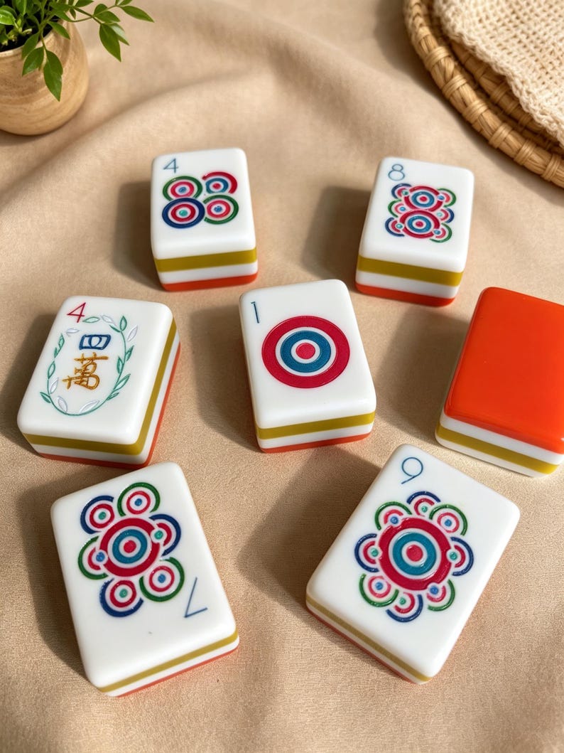 K&ouml;nnte beinhalten: Ein Set aus sieben rechteckigen Mahjong-Spielsteinen mit wei&szlig;en Oberfl&auml;chen und mehrfarbigen Seiten. Die Steine zeigen verschiedene Motive, darunter konzentrische Kreise, florale Muster, die Zahlen 4, 8, 1, 9 und das chinesische Schriftzeichen f&uuml;r zehntausend.