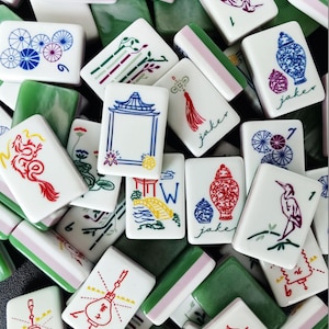May include: A collection of rectangular mahjong tiles featuring white faces with colorful illustrations of dragons, birds, vases, lanterns, and bridges, alongside green backs. Some tiles display the text jaker in a cursive script font.