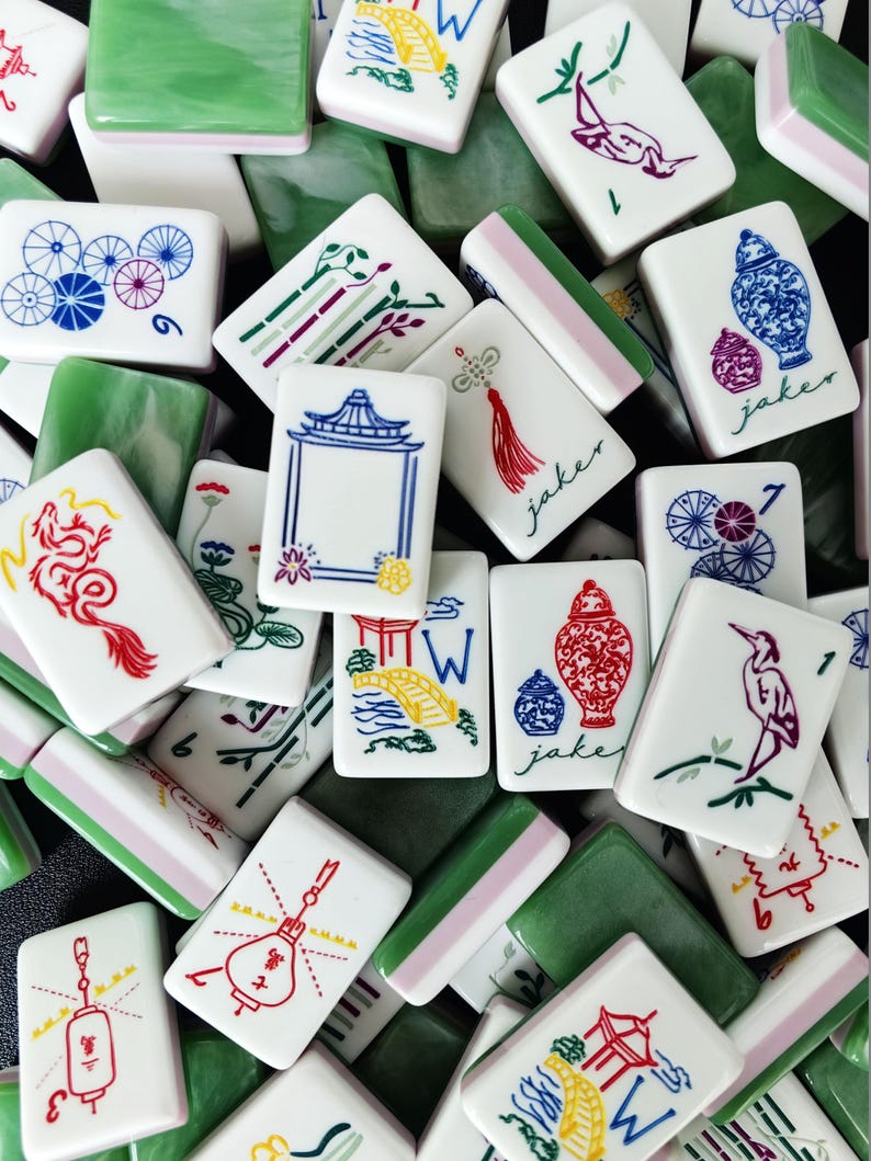 May include: A collection of rectangular mahjong tiles featuring white faces with colorful illustrations of dragons, birds, vases, lanterns, and bridges, alongside green backs. Some tiles display the text jaker in a cursive script font.