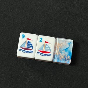 K&ouml;nnte beinhalten: Drei rechteckige Mahjong-Spielsteine, die horizontal angeordnet sind. Zwei Spielsteine zeigen eine Grafik eines Segelboots mit rotem Rumpf und blauen Segeln, markiert mit den blauen Ziffern 9 und 2. Der dritte Stein zeigt ein blau-goldenes Marmormuster.