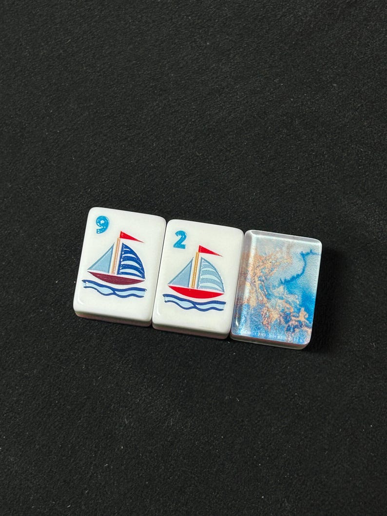 K&ouml;nnte beinhalten: Drei rechteckige Mahjong-Spielsteine, die horizontal angeordnet sind. Zwei Spielsteine zeigen eine Grafik eines Segelboots mit rotem Rumpf und blauen Segeln, markiert mit den blauen Ziffern 9 und 2. Der dritte Stein zeigt ein blau-goldenes Marmormuster.