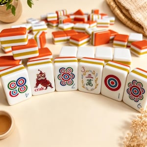 K&ouml;nnte beinhalten: Ein Set aus rechteckigen Mahjong-Spielsteinen mit wei&szlig;en, orangefarbenen und gelben horizontalen Streifen. Die Steine zeigen verschiedene bunte Grafiken, darunter einen roten L&ouml;wen, konzentrische Kreise und das chinesische Schriftzeichen f&uuml;r drei.
