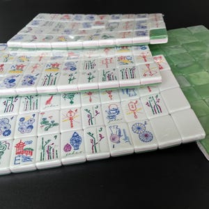 May include: A collection of white rectangular mahjong tiles featuring colorful engraved illustrations of dragons, bamboo, flowers, and lanterns, alongside a set of solid green tiles, all arranged in neat rows on a dark surface.