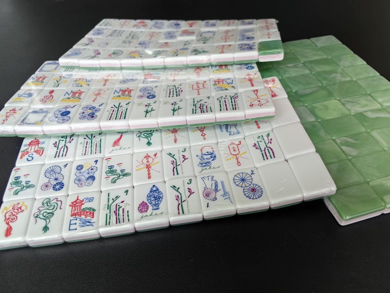 May include: A collection of white rectangular mahjong tiles featuring colorful engraved illustrations of dragons, bamboo, flowers, and lanterns, alongside a set of solid green tiles, all arranged in neat rows on a dark surface.