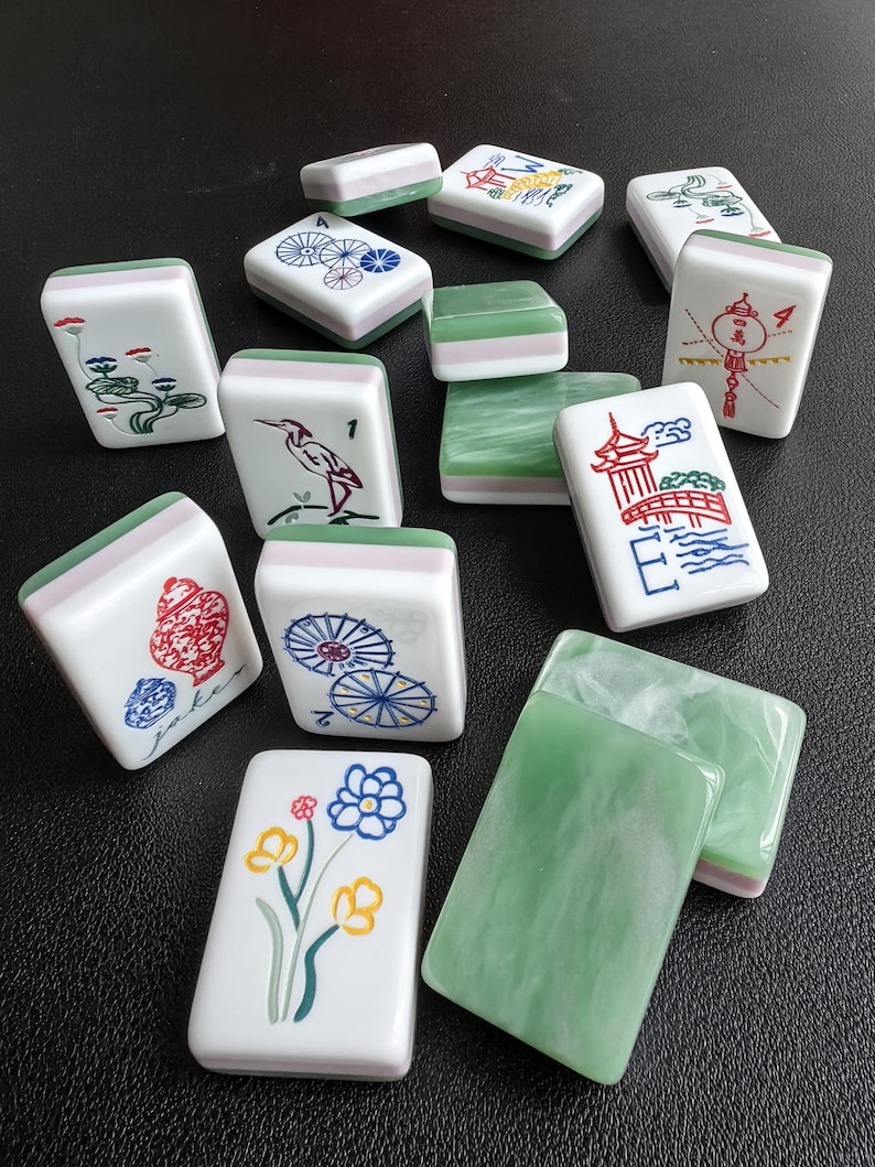 May include: A collection of rectangular Mahjong tiles featuring white faces with colorful illustrations of flowers, birds, pagodas, and umbrellas, alongside green and white marbled backs. Some tiles display the text Joker, 1, 2, and 4.