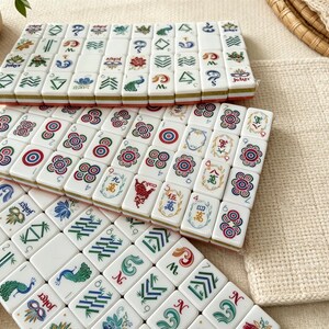 K&ouml;nnte beinhalten: Ein Set aus wei&szlig;en, rechteckigen Mahjong-Spielsteinen, die in drei Reihen angeordnet sind und bunte Illustrationen von Pfauen, Blumen, Bambus und Jokermasken zeigen. Jeder Spielstein weist verschiedene Symbole, Zahlen und Muster in Farbe auf.