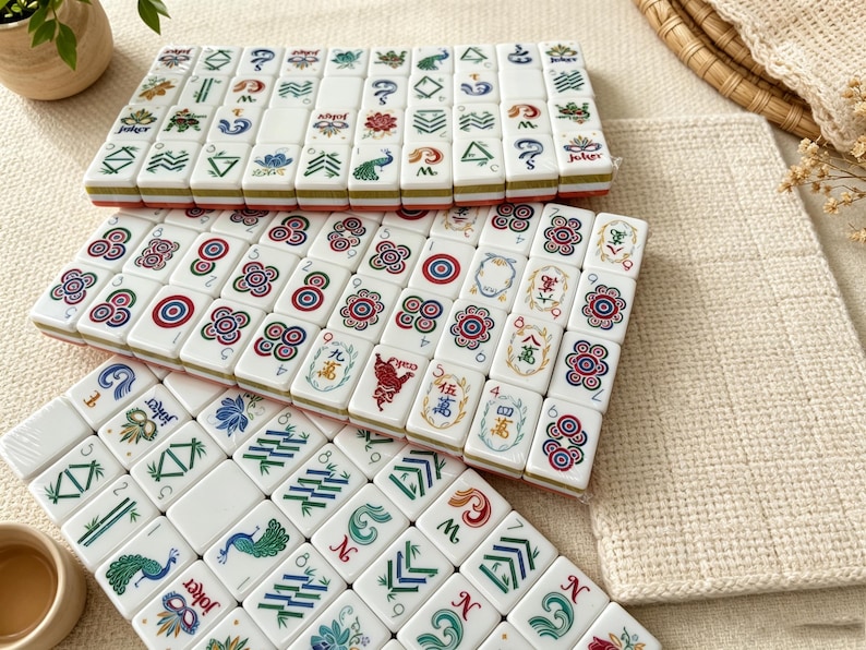 K&ouml;nnte beinhalten: Ein Set aus wei&szlig;en, rechteckigen Mahjong-Spielsteinen, die in drei Reihen angeordnet sind und bunte Illustrationen von Pfauen, Blumen, Bambus und Jokermasken zeigen. Jeder Spielstein weist verschiedene Symbole, Zahlen und Muster in Farbe auf.