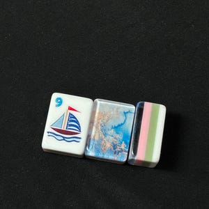 K&ouml;nnte beinhalten: Drei rechteckige Mahjong-Spielsteine, die horizontal angeordnet sind. Der erste Stein ist wei&szlig; mit einer blau-roten Segelbootgrafik und der Zahl 9. Der zweite Stein zeigt ein blau-goldenes Marmormuster. Der dritte Stein hat vertikale Streifen in Schwarz, Pink, Gr&uuml;n und Wei&szlig;.
