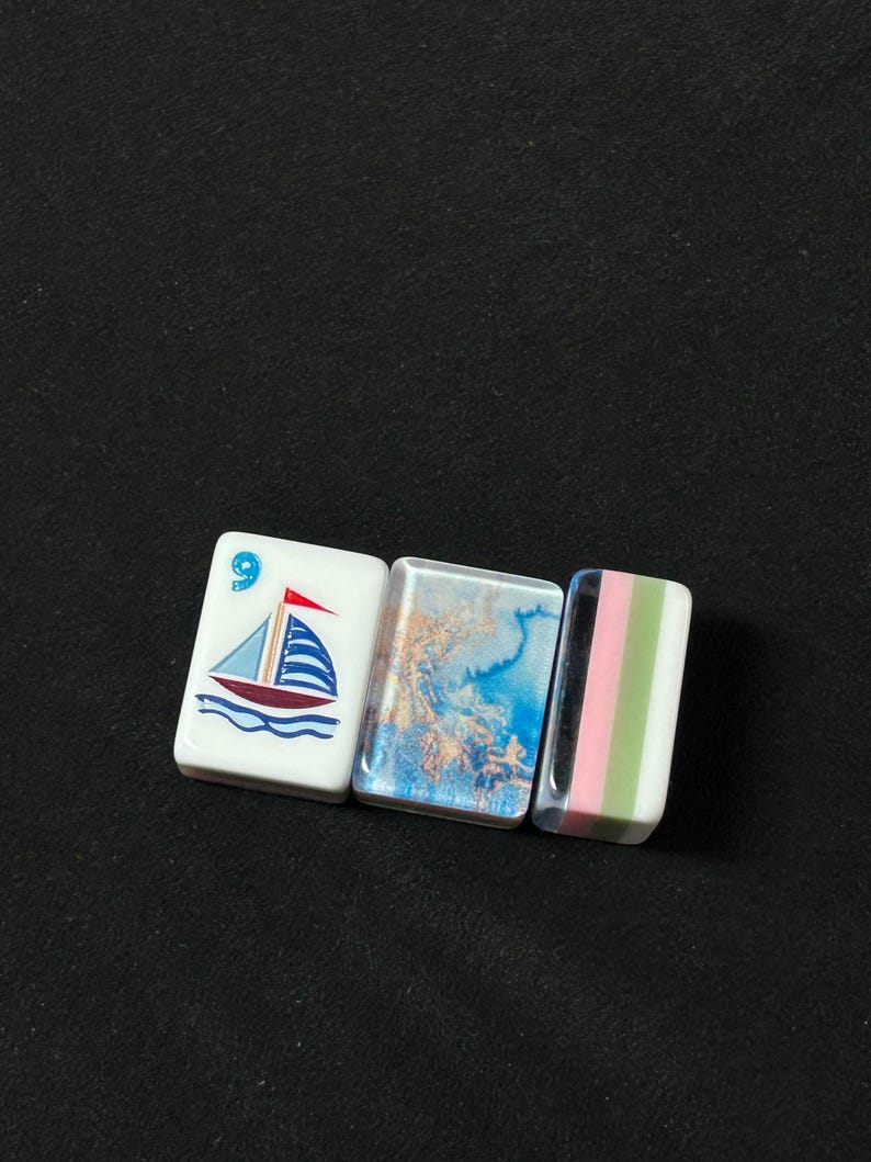 K&ouml;nnte beinhalten: Drei rechteckige Mahjong-Spielsteine, die horizontal angeordnet sind. Der erste Stein ist wei&szlig; mit einer blau-roten Segelbootgrafik und der Zahl 9. Der zweite Stein zeigt ein blau-goldenes Marmormuster. Der dritte Stein hat vertikale Streifen in Schwarz, Pink, Gr&uuml;n und Wei&szlig;.