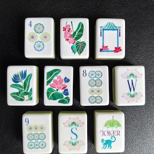 Puede incluir: Un conjunto de diez fichas de mahjong rectangulares blancas con ilustraciones bot&aacute;nicas, florales y geom&eacute;tricas coloridas. Las fichas muestran los n&uacute;meros 4, 8 y 9, las letras W y S, una pagoda, un mono con un paraguas y la palabra JOKER.