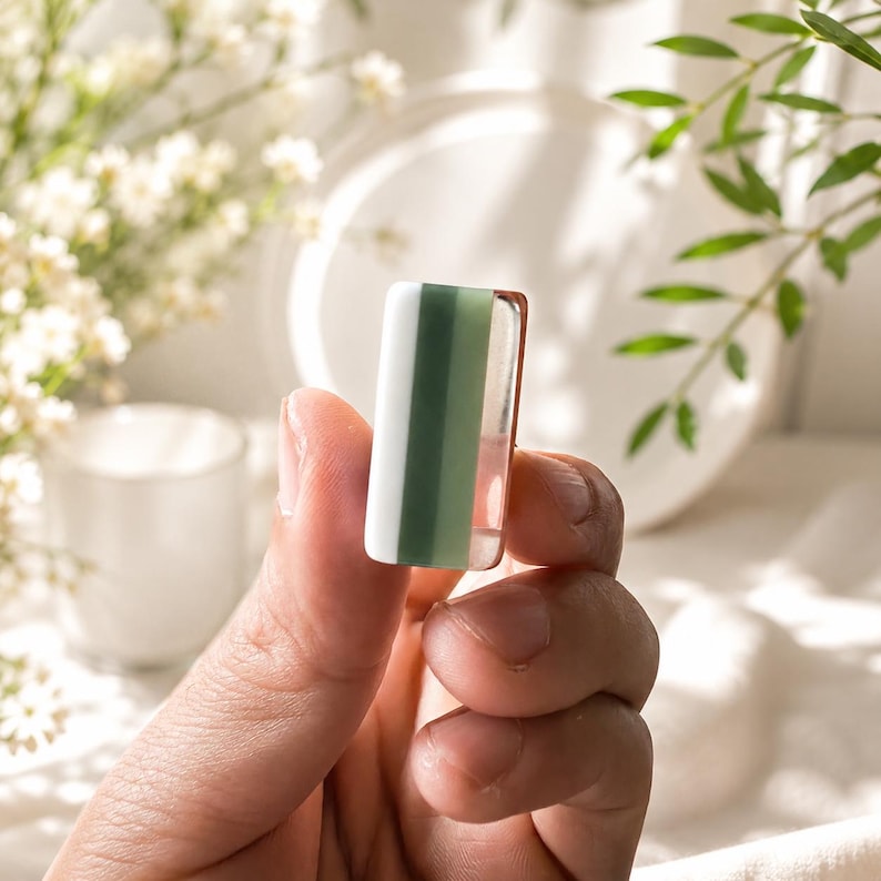 May include: A small rectangular bead featuring a vertical color-blocked design with a white section, a sage green center stripe, and a clear transparent edge. The bead has a smooth, polished finish and a uniform shape, displaying distinct color segments.