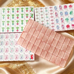 Może przedstawiać: Zestaw białych płytek do gry w Mahjong z zielonymi bambusami, czerwonymi chińskimi znakami oraz kolorowymi wzorami Jokerów i kwiatów, wraz z oddzielnym blokiem półprzezroczystych różowych płytek o gładkim, polerowanym wykończeniu.
