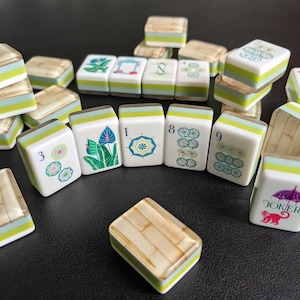 Puede incluir: Una colecci&oacute;n de fichas de Mahjong con caras blancas e ilustraciones coloridas de flores, n&uacute;meros y un mono bajo un paraguas con la etiqueta Joker. Las fichas tienen un dise&ntilde;o en capas con una franja verde claro y un reverso texturizado.