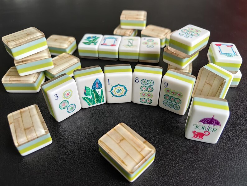 Puede incluir: Una colecci&oacute;n de fichas de Mahjong con caras blancas e ilustraciones coloridas de flores, n&uacute;meros y un mono bajo un paraguas con la etiqueta Joker. Las fichas tienen un dise&ntilde;o en capas con una franja verde claro y un reverso texturizado.