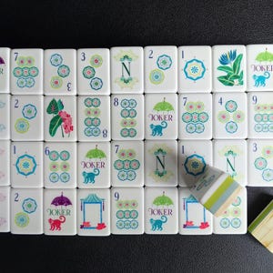 Puede incluir: Una colecci&oacute;n de fichas de mahjong rectangulares blancas con ilustraciones coloridas de paraguas, monos, flores y letras. Algunas fichas muestran n&uacute;meros del uno al nueve, mientras que otras muestran la palabra JOKER, las letras N, E, M, W o motivos florales.