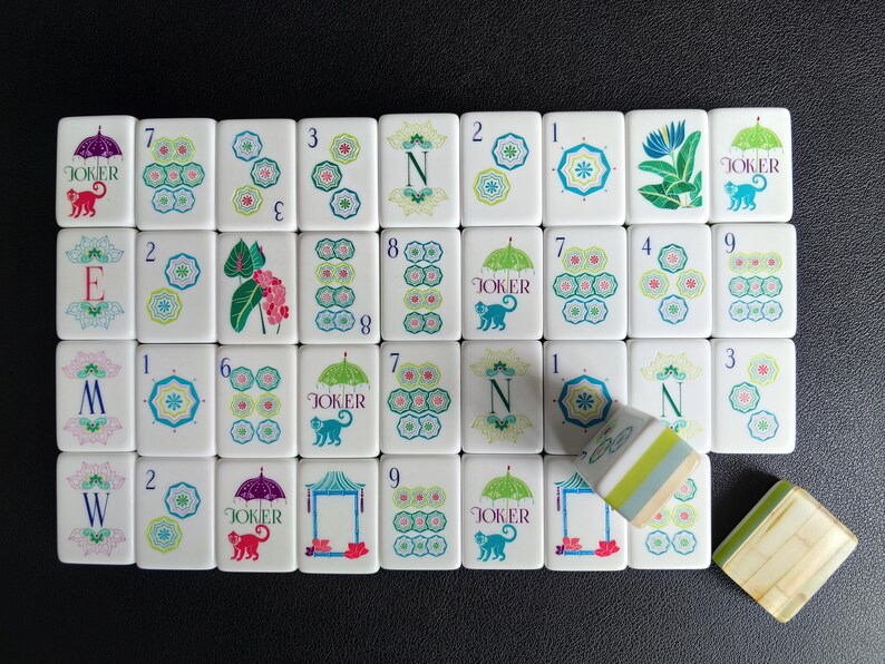 Puede incluir: Una colecci&oacute;n de fichas de mahjong rectangulares blancas con ilustraciones coloridas de paraguas, monos, flores y letras. Algunas fichas muestran n&uacute;meros del uno al nueve, mientras que otras muestran la palabra JOKER, las letras N, E, M, W o motivos florales.