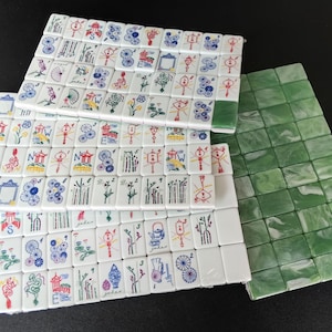 May include: A collection of rectangular Mahjong tiles featuring white faces with colorful engraved illustrations of dragons, flowers, bamboo, and directional letters, alongside a set of solid green marbled tiles with blank faces.