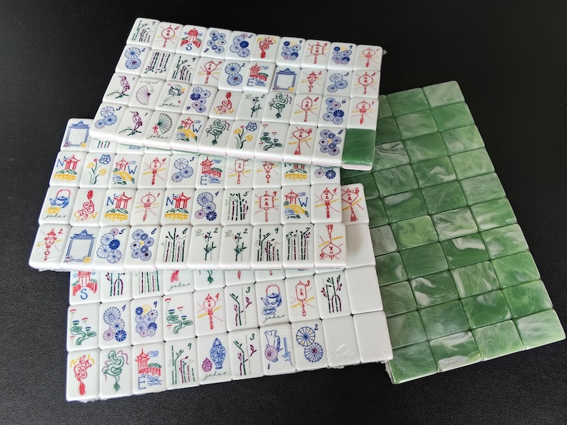 May include: A collection of rectangular Mahjong tiles featuring white faces with colorful engraved illustrations of dragons, flowers, bamboo, and directional letters, alongside a set of solid green marbled tiles with blank faces.