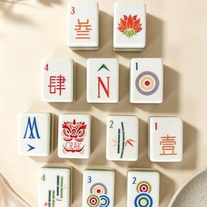 May include: A collection of eleven white rectangular Mahjong tiles arranged in a pyramid formation. Each tile features distinct colorful illustrations, including Chinese characters, a lotus flower, a lion head with the text CRAK, and bamboo.