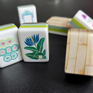 Puede incluir: Una colecci&oacute;n de fichas de Mahjong rectangulares con caras blancas e ilustraciones coloridas que incluyen una flor azul, un n&uacute;mero siete con patrones florales y otros s&iacute;mbolos. Algunas fichas muestran un perfil lateral verde claro.