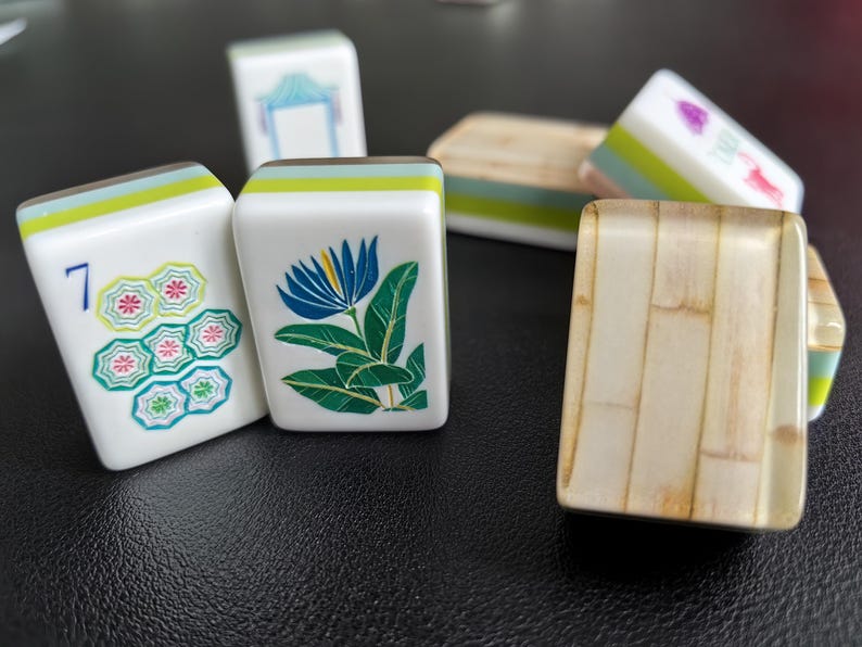 Puede incluir: Una colecci&oacute;n de fichas de Mahjong rectangulares con caras blancas e ilustraciones coloridas que incluyen una flor azul, un n&uacute;mero siete con patrones florales y otros s&iacute;mbolos. Algunas fichas muestran un perfil lateral verde claro.