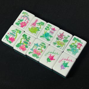 Op de afbeelding: Een set van negen rechthoekige mahjongtegels met witte voorzijden. Zeven tegels bevatten kleurrijke illustraties van pagodes, lotusbloemen en bomen in roze, groen en blauw. Twee tegels tonen het woord joker in cursief schrift boven een narrenkap.