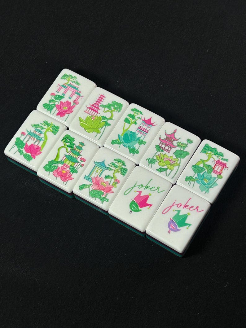 Op de afbeelding: Een set van negen rechthoekige mahjongtegels met witte voorzijden. Zeven tegels bevatten kleurrijke illustraties van pagodes, lotusbloemen en bomen in roze, groen en blauw. Twee tegels tonen het woord joker in cursief schrift boven een narrenkap.