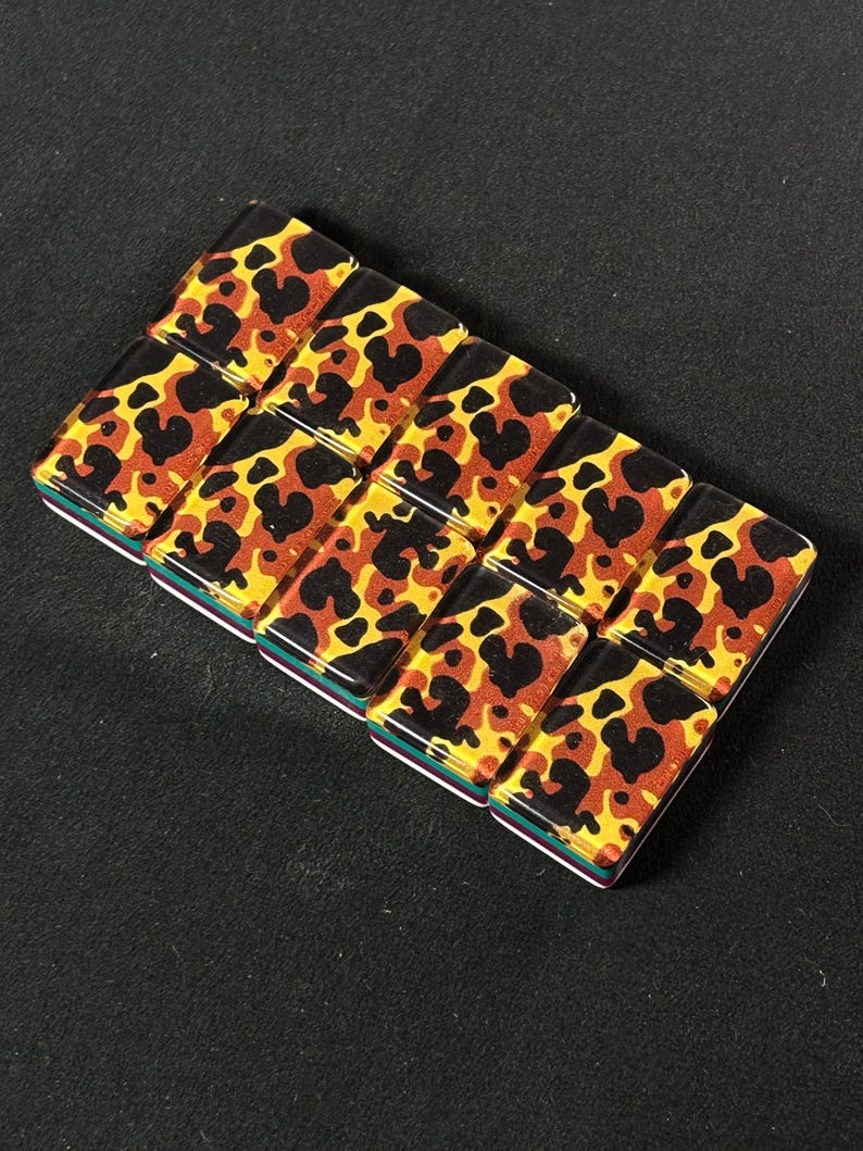 Op de afbeelding: Een rechthoekige armband bestaande uit tien kleine, gladde, rechthoekige tegeltjes gerangschikt in twee rijen. Elk tegeltje heeft een glanzende, doorschijnende afwerking met een luipaardprint in tinten zwart, bruin en goudgeel.