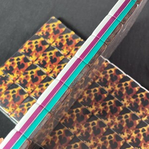 Op de afbeelding: Twee rechthoekige tegels met een herhalend luipaardpatroon in tinten zwart, bruin en geel. De zijkanten van de tegels zijn gelaagd met opvallende strepen in wit, magenta en turquoise, wat zorgt voor een kleurrijke rand.