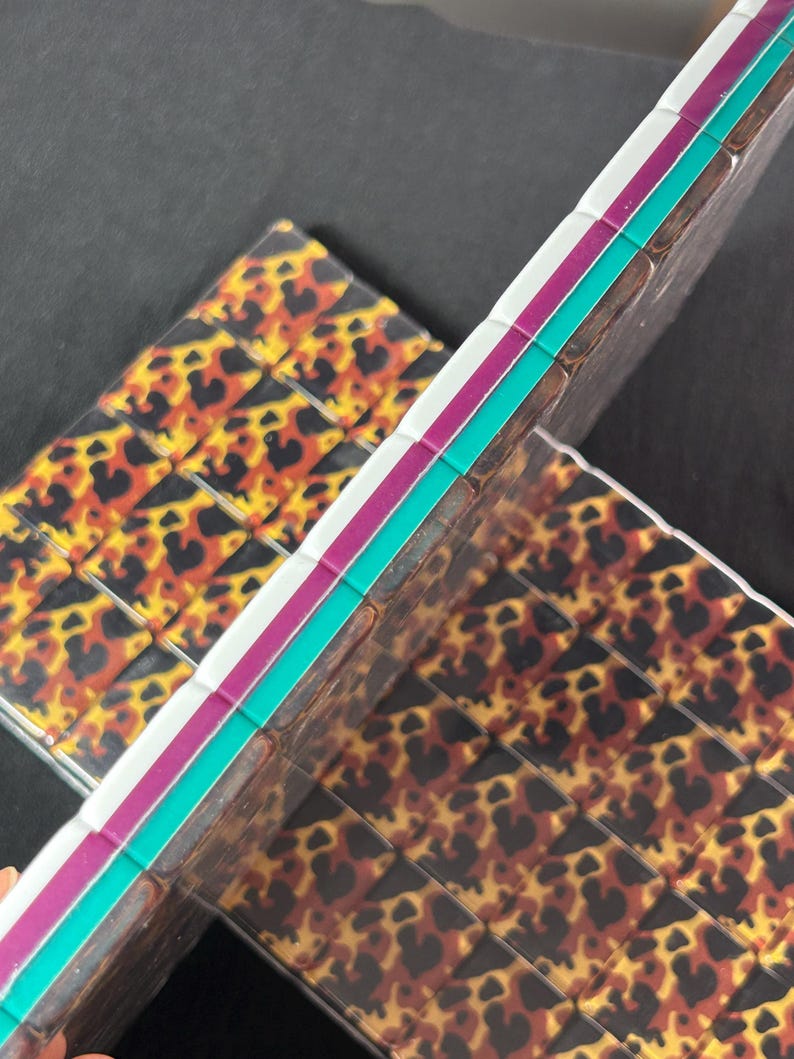 Op de afbeelding: Twee rechthoekige tegels met een herhalend luipaardpatroon in tinten zwart, bruin en geel. De zijkanten van de tegels zijn gelaagd met opvallende strepen in wit, magenta en turquoise, wat zorgt voor een kleurrijke rand.