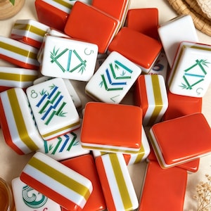 K&ouml;nnte beinhalten: Eine Sammlung rechteckiger Mahjong-Spielsteine mit einer Mischung aus einfarbigen orangefarbenen R&uuml;ckseiten und wei&szlig;en Vorderseiten mit gr&uuml;nen und blauen Bambusmustern, Zahlensymbolen und dekorativen Designs. Die Steine liegen verstreut.