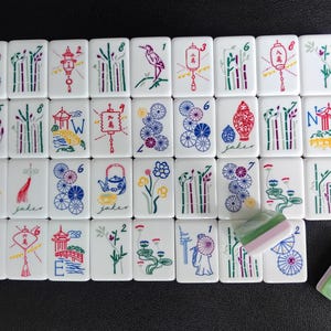 May include: A collection of white rectangular mahjong tiles featuring colorful illustrations of bamboo, lanterns, flowers, fans, and pagodas. Some tiles display numbers and letters, while one tile is partially covered by a green and white piece.