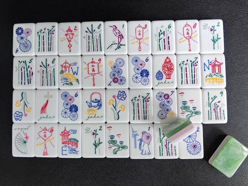May include: A collection of white rectangular mahjong tiles featuring colorful illustrations of bamboo, lanterns, flowers, fans, and pagodas. Some tiles display numbers and letters, while one tile is partially covered by a green and white piece.