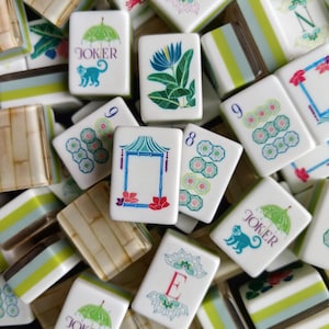 Puede incluir: Una colecci&oacute;n de fichas de mahjong blancas con ilustraciones coloridas que incluyen una flor azul, una pagoda, paraguas verdes con monos y las letras W, N y E. Algunas fichas muestran patrones num&eacute;ricos, mientras otras tienen textura de bamb&uacute;.