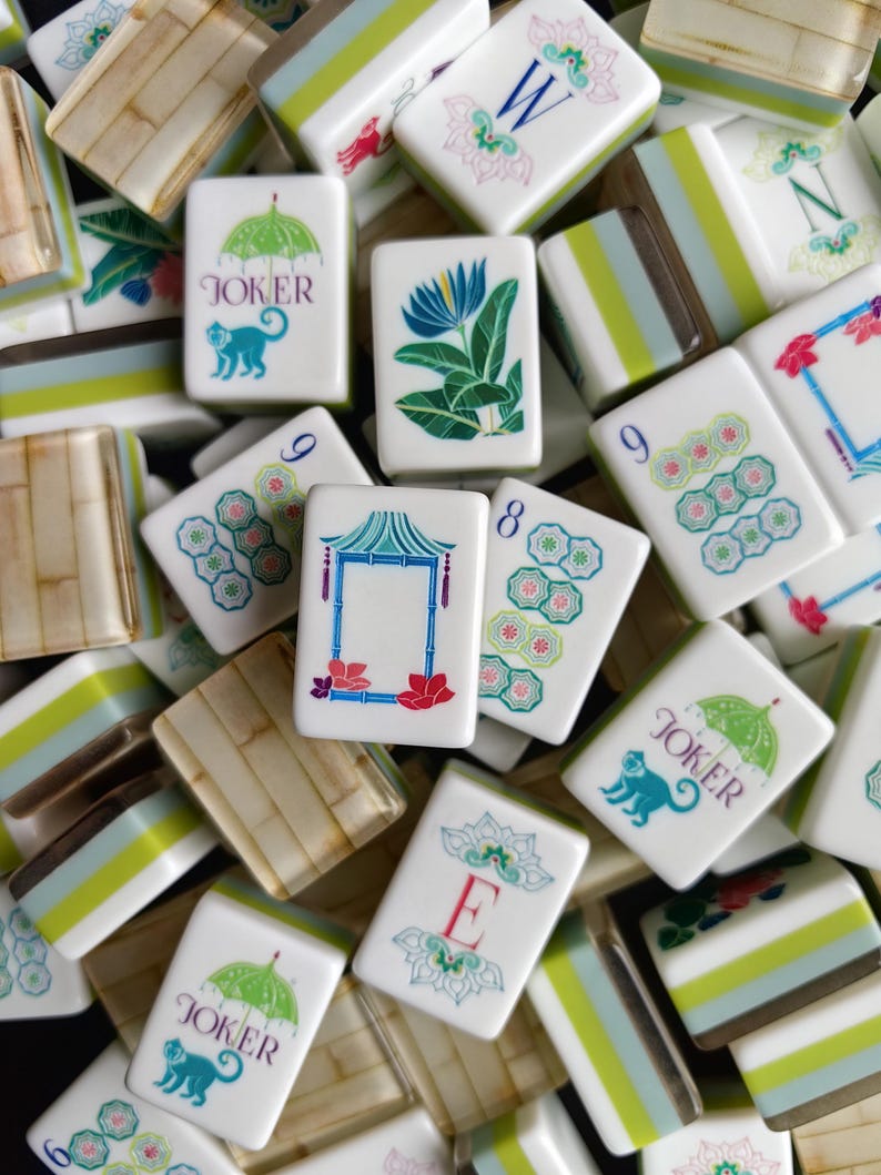 Puede incluir: Una colecci&oacute;n de fichas de mahjong blancas con ilustraciones coloridas que incluyen una flor azul, una pagoda, paraguas verdes con monos y las letras W, N y E. Algunas fichas muestran patrones num&eacute;ricos, mientras otras tienen textura de bamb&uacute;.